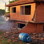 Residência em Ceilândia danificada por explosão de botijões de gás, com botijões espalhados e viatura de bombeiros ao fundo.