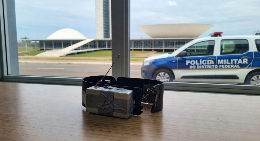 Tornozeleira eletrônica burlada com dispositivo em delegacia no DF, viatura policial ao fundo.