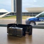 Tornozeleira eletrônica burlada com dispositivo em delegacia no DF, viatura policial ao fundo.