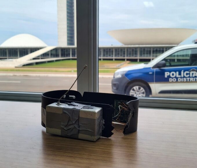 Tornozeleira eletrônica burlada com dispositivo em delegacia no DF, viatura policial ao fundo.