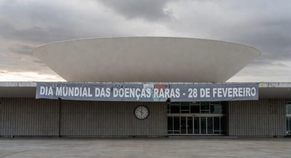 Edifício da CLDF em Brasília com banner atrasado sobre Dia das Doenças Raras, expondo negligência no apoio a pacientes.