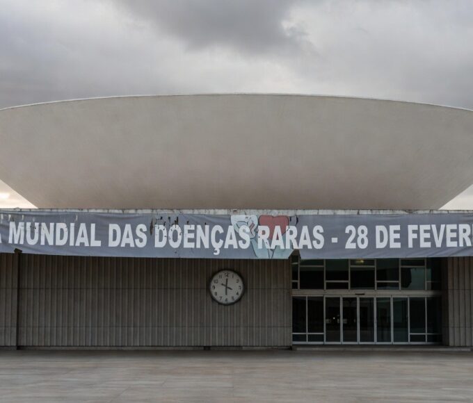 Edifício da CLDF em Brasília com banner atrasado sobre Dia das Doenças Raras, expondo negligência no apoio a pacientes.