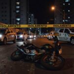 Cena de crime em rua de Taguatinga com viatura policial, fita de isolamento e motocicleta caída, representando duplo homicídio.